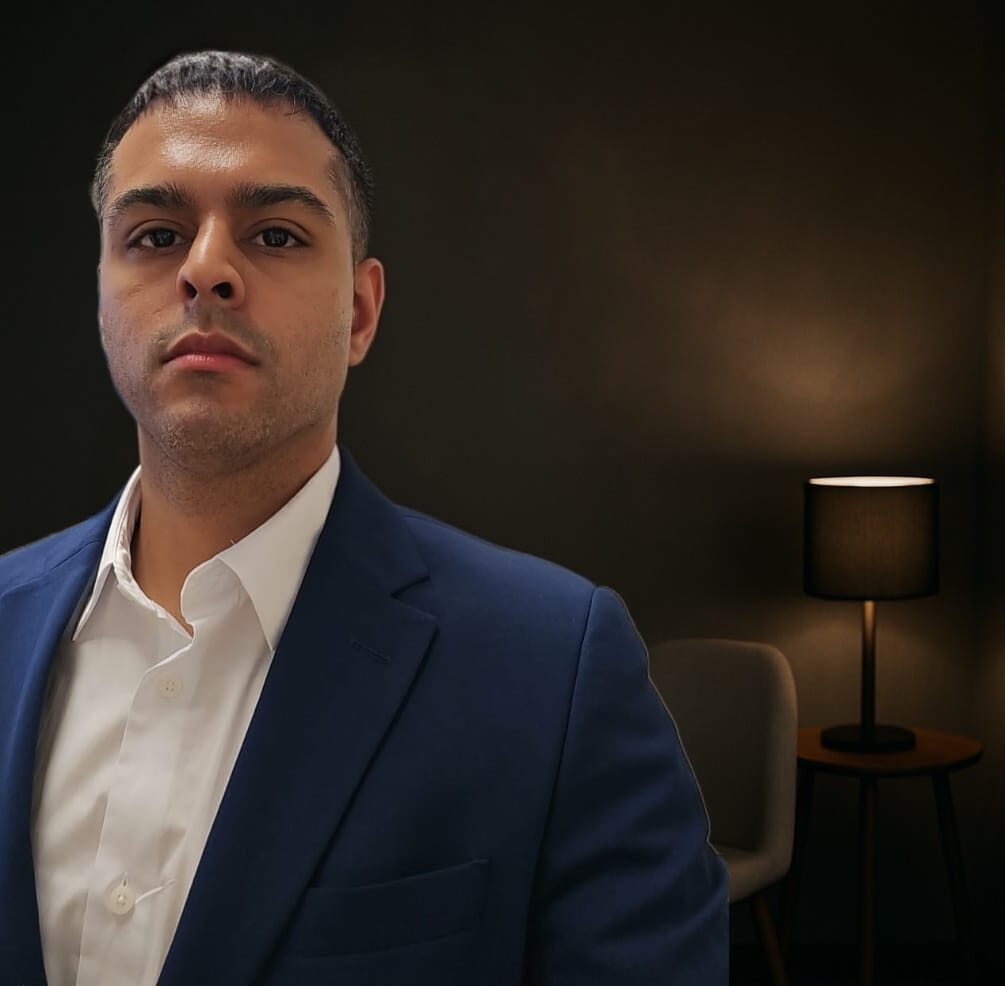 A professional man in a navy blue suit standing in a dimly lit room with a chair and lamp behind him, symbolizing focus and clarity during a personal coaching session.